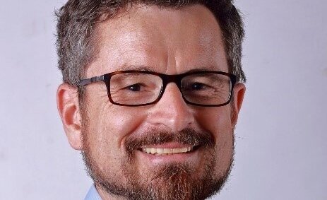 Porträt eines lächelnden Mannes mit Brille und Bart vor neutralem Hintergrund.