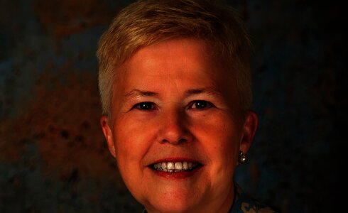 Eine lächelnde Frau mit kurzen, blonden Haaren vor einem blauen Hintergrund.