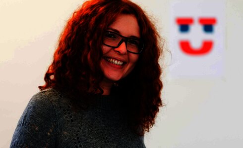 Lächelnde Frau mit lockigem, rotem Haar und Brille in grauem Pullover.