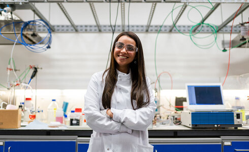 Eine Wissenschaftlerin in einem Labor mit Schutzbrille und Laborkittel lächelt.