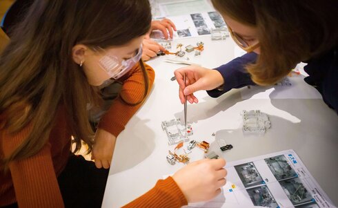 Zwei Personen arbeiten gemeinsam mit elektrischen Bauteilen und Anleitungen am Tisch.