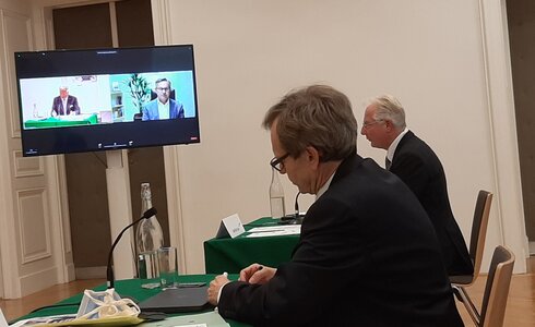 Zwei Personen in Anzügen nehmen an einer Videokonferenz in einem Besprechungsraum teil.