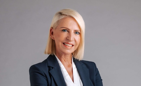 Professionelle Frau in Anzug, lächelnd, mit verschränkten Armen vor neutralem Hintergrund.