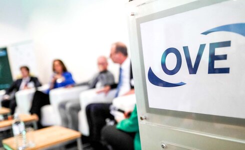 Logo der OVE vor einer Paneldiskussion mit mehreren Teilnehmern im Hintergrund.
