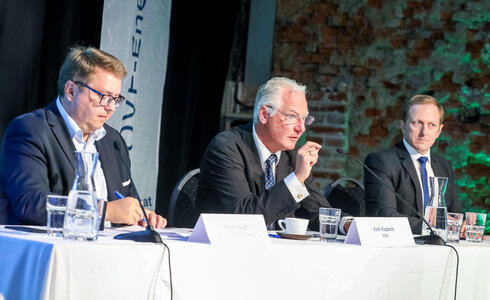 Drei Männer in Anzügen an einem Tisch während einer Diskussion oder Präsentation.