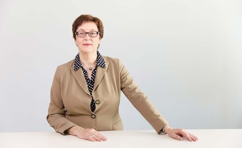Eine ältere Frau in einem blazer sitzt an einem Tisch und schaut freundlich.