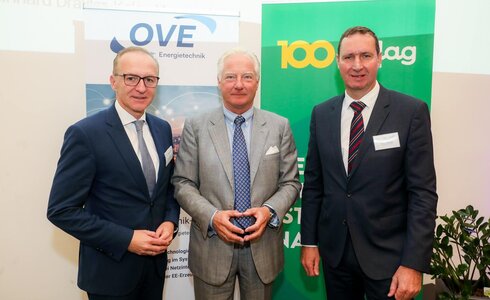 Drei Männer posieren zusammen bei einer Veranstaltung mit Bannern im Hintergrund.