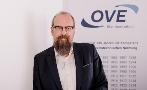 Mann mit Brille und Bart vor Hintergrund mit OVE-Logo und Text zur Standardisierung.