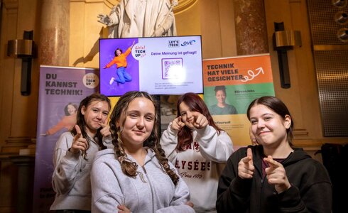 Vier junge Frauen posieren vor einem Bildschirm mit dem Motto „Deine Meinung ist gefragt“.