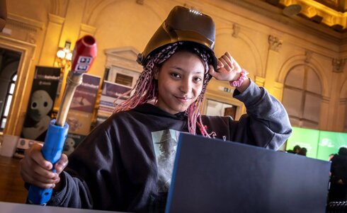 Junge Frau mit Schweißgerät und Schutzhelm sitzt vor einem Laptop.