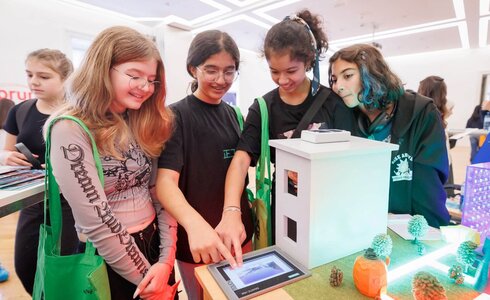 Vier Mädchen betrachten ein Modellhaus und interagieren mit einem Tablet.