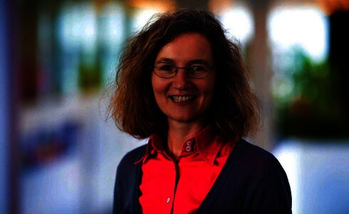 Lächelnde Frau mit Brille, trägt ein pinkes Hemd und einen grauen Pullover.