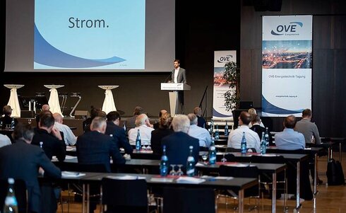 Mann spricht auf Konferenz, Hintergrund zeigt Präsentation über Strom.