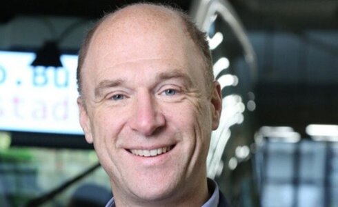 Ein lächelnder Mann mit kurzen Haaren und blauer Jacke vor einem unscharfen Hintergrund.