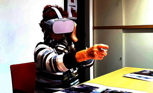 Eine ältere Frau trägt eine VR-Brille und interagiert mit Bildern am Tisch.