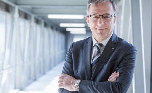 Mann in Anzug steht mit verschränkten Armen und lächelt selbstbewusst.