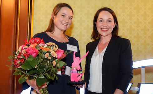 Zwei Frauen halten eine Trophäe und einen Blumenstrauß bei einer Auszeichnung.