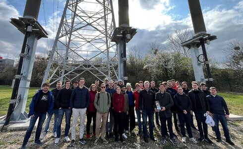 Gruppe von Personen steht vor einem hohen Strommasten im Freien.