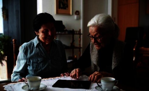 Zwei Frauen sitzen gemeinsam am Tisch und schauen auf ein Tablet.