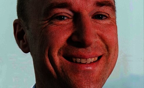 Ein Mann lächelt freundlich in Anzug und Krawatte vor unscharfem Hintergrund.