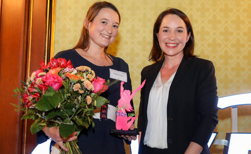 Zwei Frauen präsentieren eine Auszeichnung und einen Blumenstrauß bei einer Preisverleihung.