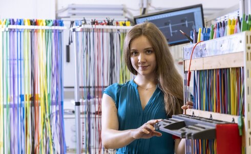 Eine junge Frau in blauer Bluse steht vor farbigen Kabelsträngen.