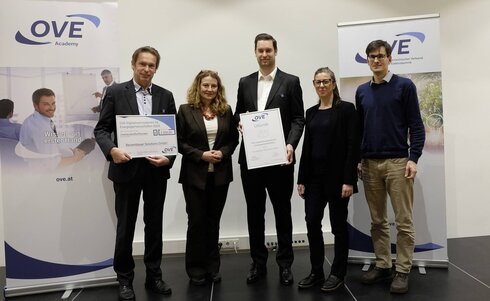 Zwei Personen halten Urkunden, umgeben von drei weiteren Menschen in formeller Kleidung.