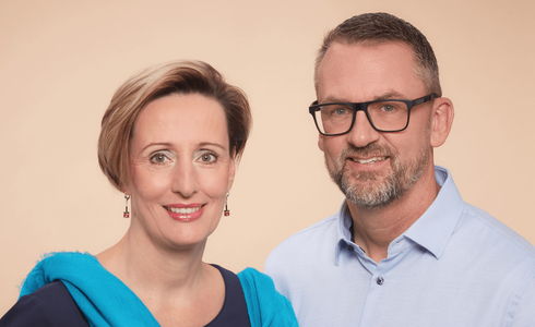 Ein Paar posiert freundlich vor einem neutralen, hellen Hintergrund.