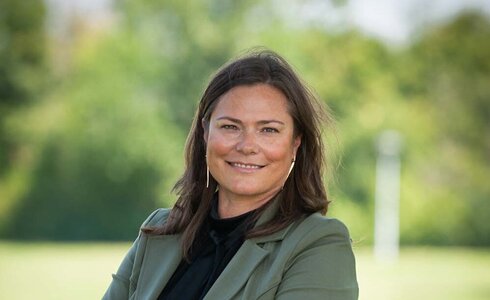 Eine Frau in einem blauen Oberteil und grünem Blazer steht lächelnd im Freien.