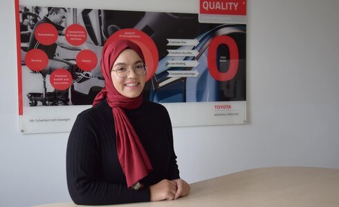 Junge Frau mit Kopftuch lächelt vor einem Qualitätsschild von Toyota.